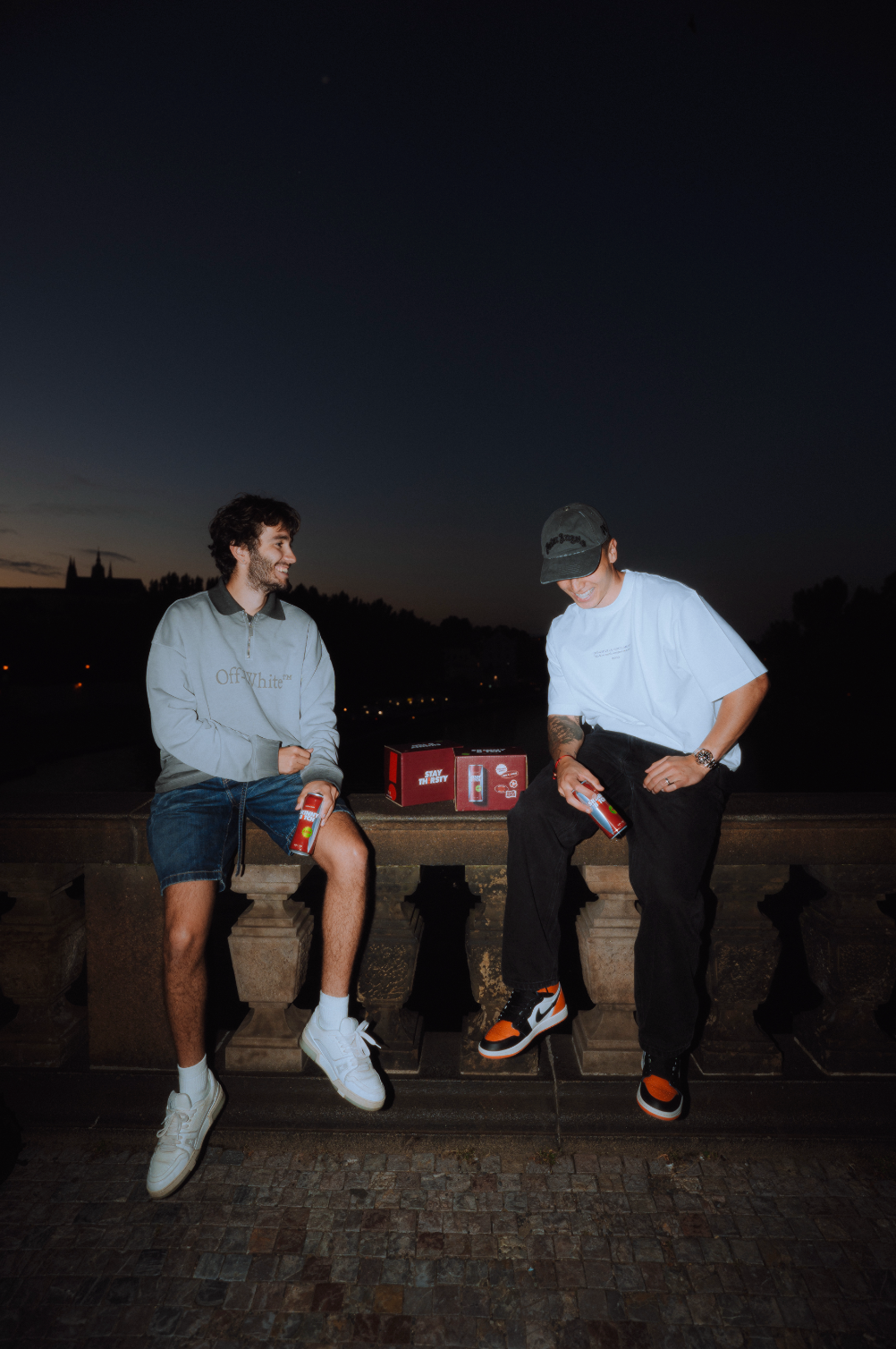 Two friends on a Prague bridge at dusk with cans and six-packs